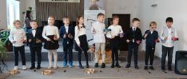 Uczniowie klasy 1 cyklu 6-letniego występują na scenie. Dzieci stoją w rzędzie i śpiewają piosenkę.