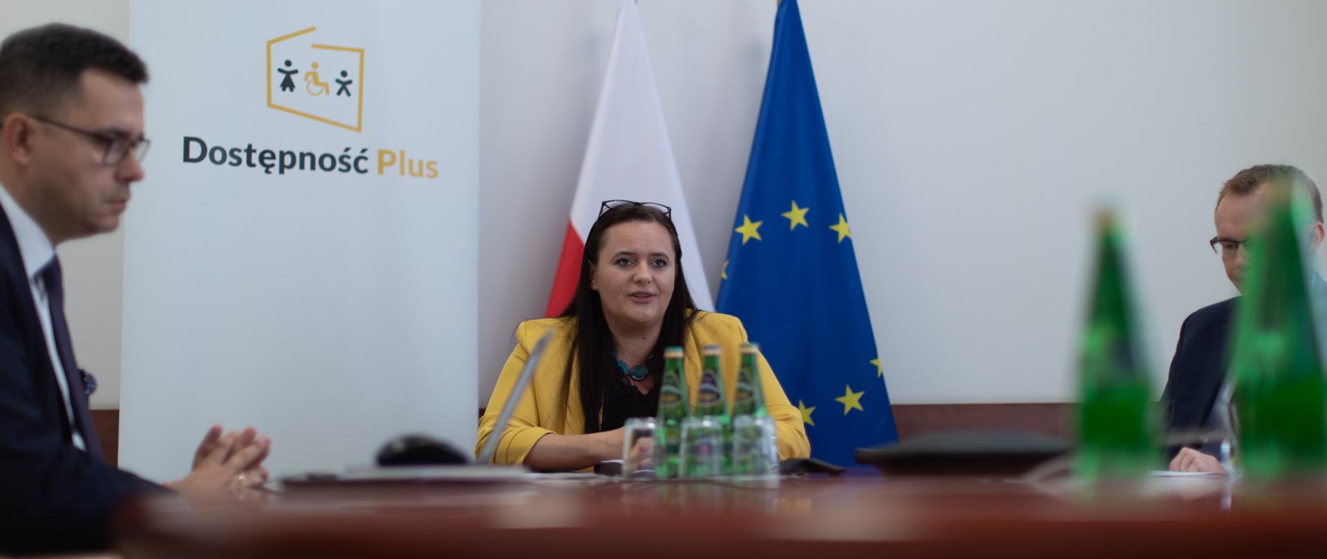 W pomieszczeniu przy stole siedzą trzy osoby. Wśród nich obok białego Roll-upu z napisem Dostępność Plus siedzi minister M. Jarosińska-Jedynak.