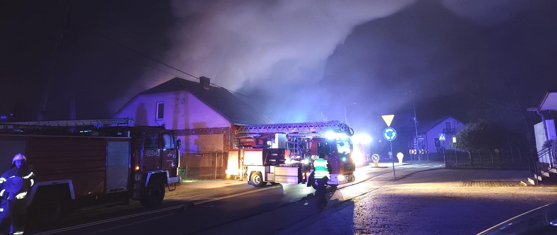 Przedstawia akcję gaśniczą w porze nocnej. Na ulicy przed budynkiem stoją wozy strażackie z włączonymi światłami ostrzegawczymi, a w górę unosi się gęsty, oświetlony dym. Widoczni są strażacy w pełnym ekwipunku zabezpieczający miejsce zdarzenia.