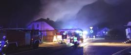 Przedstawia akcję gaśniczą w porze nocnej. Na ulicy przed budynkiem stoją wozy strażackie z włączonymi światłami ostrzegawczymi, a w górę unosi się gęsty, oświetlony dym. Widoczni są strażacy w pełnym ekwipunku zabezpieczający miejsce zdarzenia.
