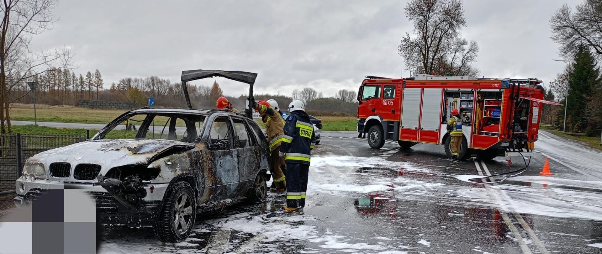 Na 1 planie po lewej stronie stoi spalony samochód a przy nim strażacy, na ulicy płynie reszta piany gaśniczej, w tle samochód strażacki, a przy nim strażak. W oddali drzewa