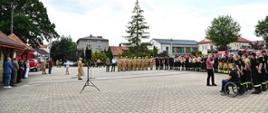 Na płycie rynku wyłożonej kostką stoją strażacy formując szyk w kształcie litery C przed nimi prezydium z zaproszonymi gośćmi. W tle budynki oraz drzewa