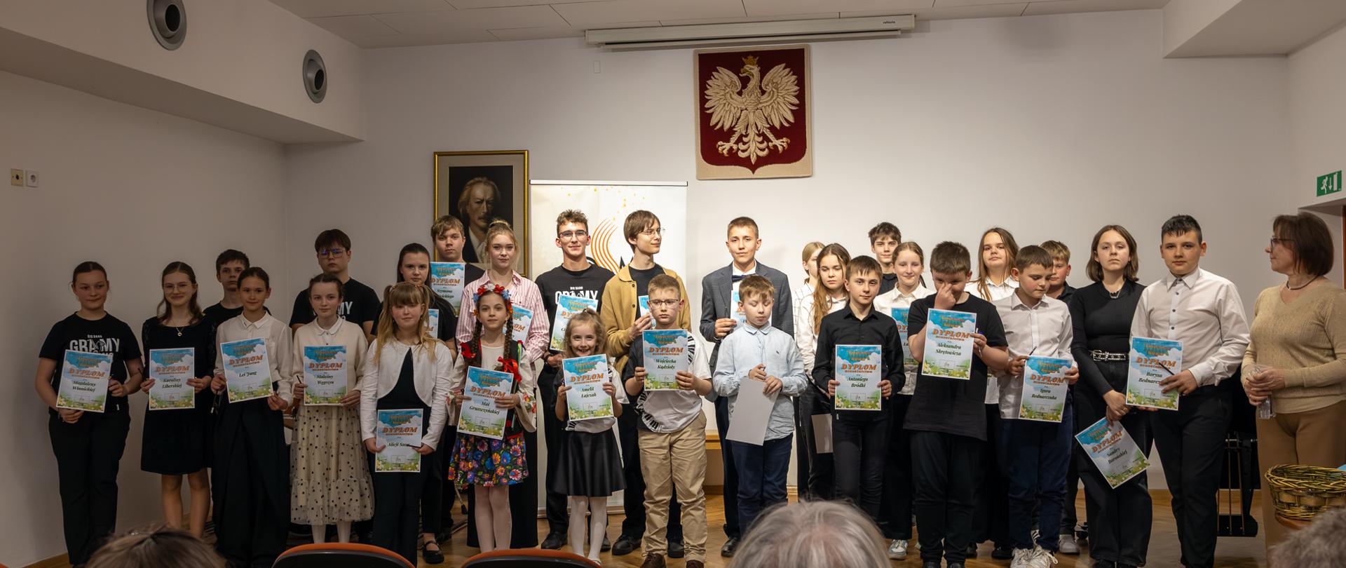 Uczestnicy szkolnego konkursu na scenie szkoły muzycznej, wszyscy trzymają w rękach dyplomy uczestnictwa. 