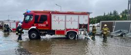 Usuwanie skutków intensywnych opadów deszczu. Zdjęcie przedstawia podtopiony parking i plac magazynowy marketu budowlanego. Na placu znajdują się dwa wozy pożarnicze i 5 strażaków uczestniczących w akcji. Do jednego z samochodów podlączone są węże.