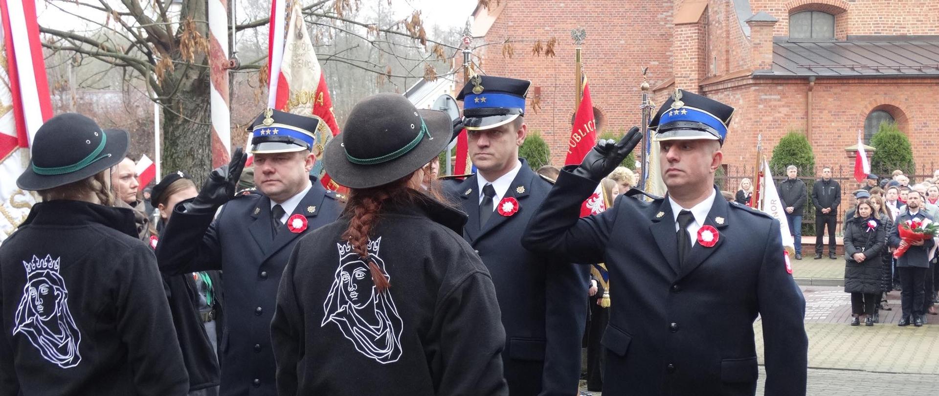 Na zdjęciu widać strażaków w galowych mundurach oraz osoby związane ze strażą pożarną, uczestniczących w uroczystości z okazji 11 listopada. Grupa oficerów i towarzysząca im osoba w cywilnym stroju, na którym widoczne jest haftowane godło, salutuje, stojąc przed budynkiem z czerwonej cegły. W tle zgromadzeni są inni uczestnicy i poczty sztandarowe, co podkreśla podniosły charakter składania hołdu w Narodowe Święto Niepodległości.