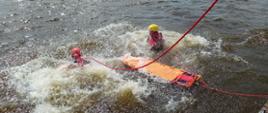 Zdjęcie przedstawia dwóch strażaków w skafandrach ratowniczych, kaskach i kamizelkach asekuracyjnych płynących do osoby topiącej się. Strażacy płyną z noszami typu deska o dodatniej pływalności jako środkiem ewakuacji poszkodowanego 