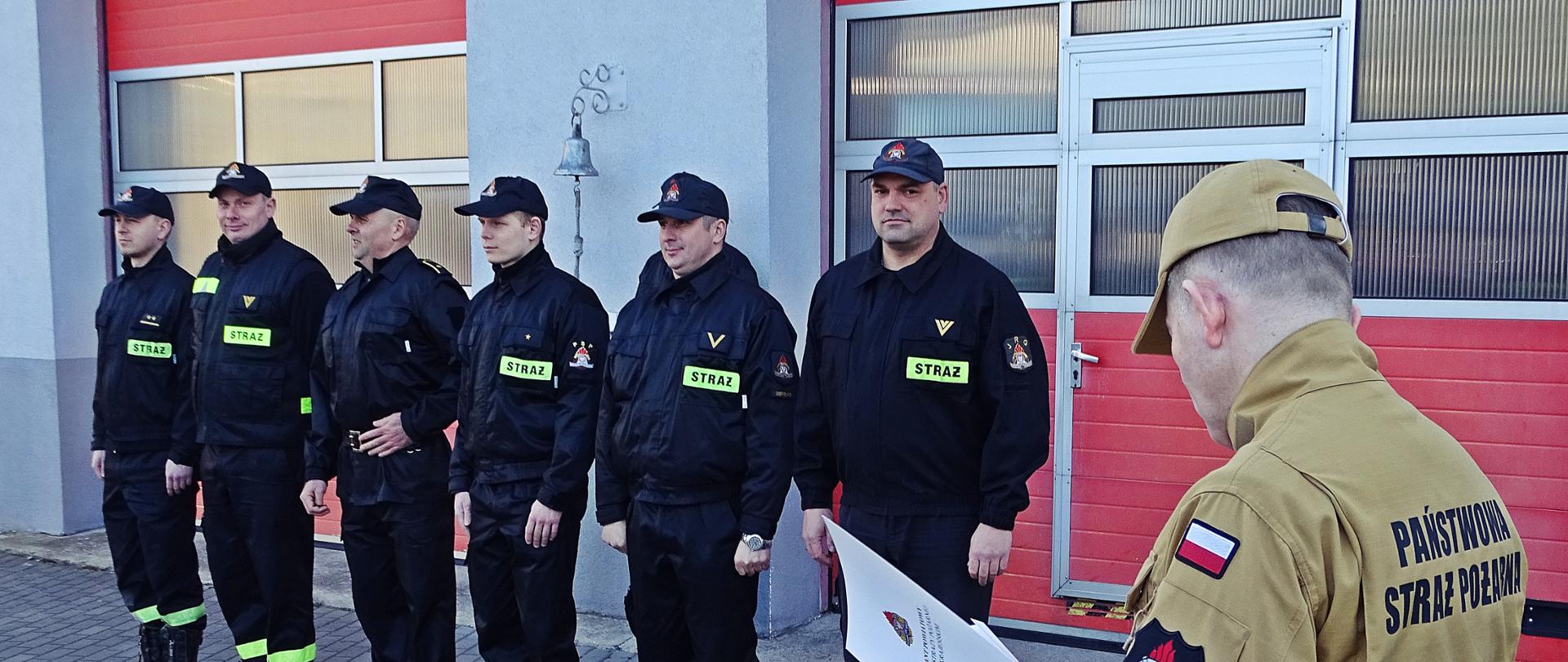 Zdjęcie wykonano w ciągu dnia na placu Komendy Powiatowej Państwowej Straży Pożarnej w Sępólnie Krajeńskim. Komendant Powiatowy Państwowej Straży Pożarnej starszy brygadier Maciej Suchomski w mundurze służbowym(po prawej) odczytuje pismo służbowe. W tle strażacy zmiany służbowej stoją w szeregu.