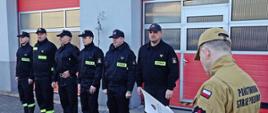 Zdjęcie wykonano w ciągu dnia na placu Komendy Powiatowej Państwowej Straży Pożarnej w Sępólnie Krajeńskim. Komendant Powiatowy Państwowej Straży Pożarnej starszy brygadier Maciej Suchomski w mundurze służbowym(po prawej) odczytuje pismo służbowe. W tle strażacy zmiany służbowej stoją w szeregu.