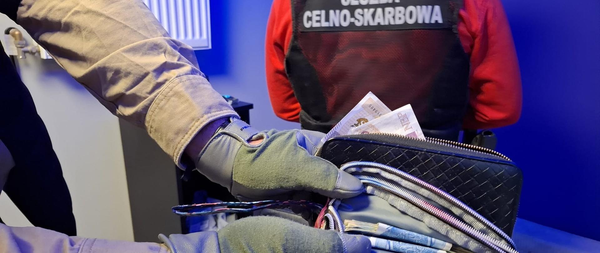 pomieszczenie, plecy mężczyzny w czarnej kamizelce z napisem służba celno-skarbowa, ręce drugiego mężczyzny trzymające dwa portfele pełne pieniędzy