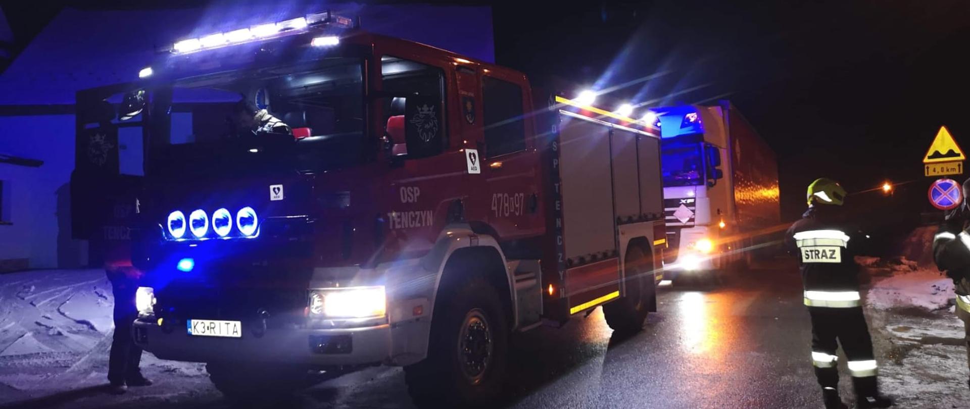 Zdjęcie pokazuje samochód strażacki OSP Tenczyn oraz widoczny za nim samochód ciężarowy z naczepą, który został odholowany w bezpieczne miejsce. Z prawej strony widoczny strażak w umundurowaniu specjalnym.