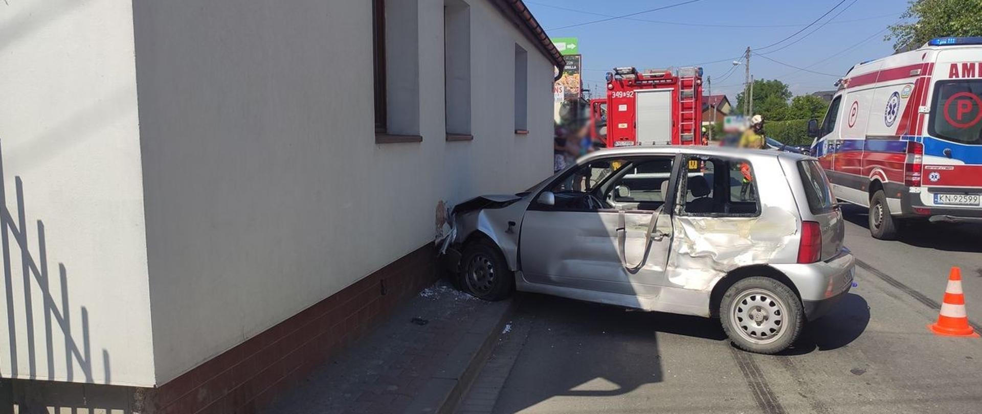 Samochód osobowy przodem uderzył w ścianę budynku. Zniszczony lewy bok. W tle samochód pożarniczy oraz karetka Pogotowia Ratunkowego. 
