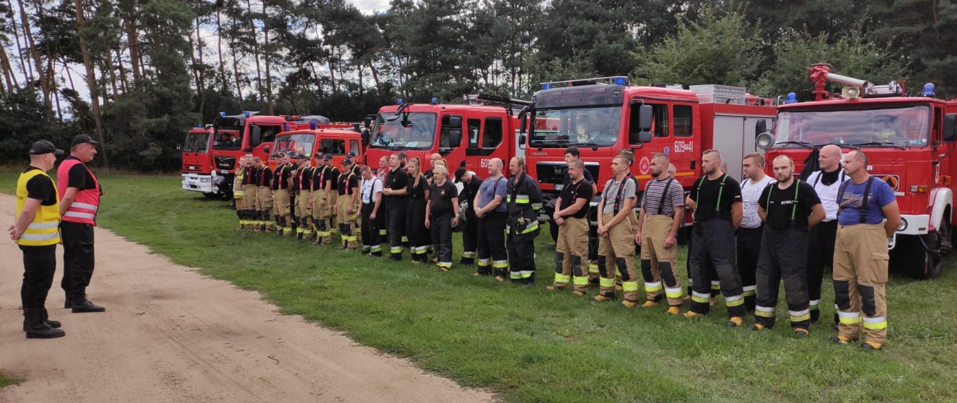 Na zdjęciu odprawa w ramach ćwiczeń 2 strażaków przekazuje informacje stojącym ratownikom z wszystkich OSP z terenu gminy Książ Wlkp. którzy stoją przed pojazdami ratowniczo - gaśniczymi 