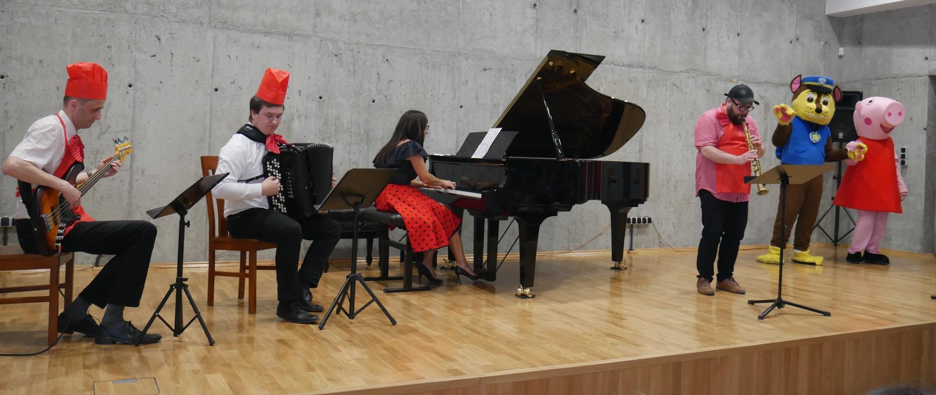 Grupa nauczycieli gra koncert na scenie sali koncertowej, ubrani są w śmieszne stroje, na scenie stoją osoby w strojach postaci z bajek