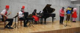 Grupa nauczycieli gra koncert na scenie sali koncertowej, ubrani są w śmieszne stroje, na scenie stoją osoby w strojach postaci z bajek
