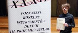 Zdjęcie przedstawia laureata Piotra Szkudlarka stojącego na scenie z dyplomem.