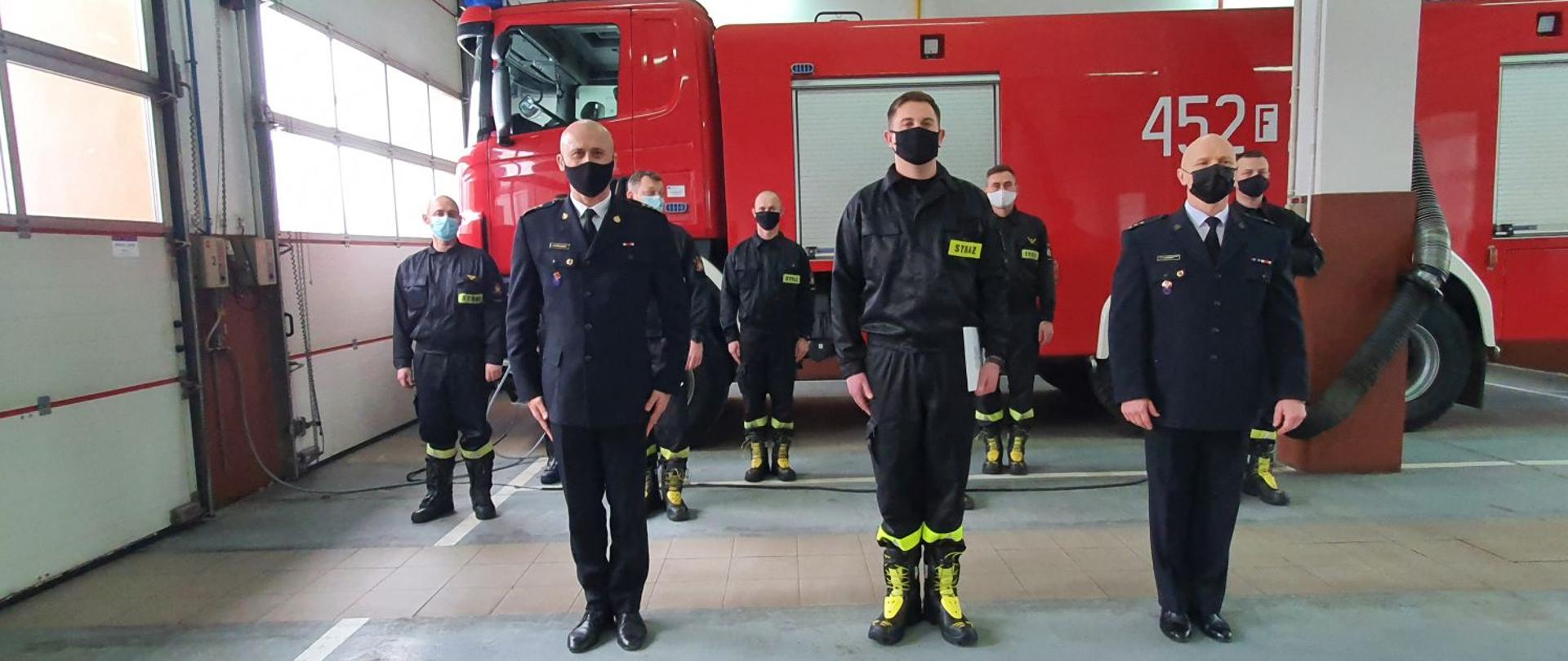 Na pierwszym planie widoczny jest Komendant Powiatowy PSP, strażak składający ślubowanie i Dowódca JRG. W drugim rzędzie obecni są strażacy ze zmiany służbowej. W tle widoczny jest samochód strażacki. 