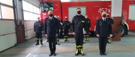 Na pierwszym planie widoczny jest Komendant Powiatowy PSP, strażak składający ślubowanie i Dowódca JRG. W drugim rzędzie obecni są strażacy ze zmiany służbowej. W tle widoczny jest samochód strażacki. 