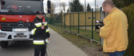 Przygotowywanie spotu edukacyjnego. Jedna z ulic w Dębnie Polskim. Strażak na tle wozu ratowniczo-gaśniczego, przed kamerą przedstawia swoją kwestię. W ręku trzyma mikrofon. Kilka metrów przed nim, na chodniku stoi operator kamery. W tle płot i budynki.