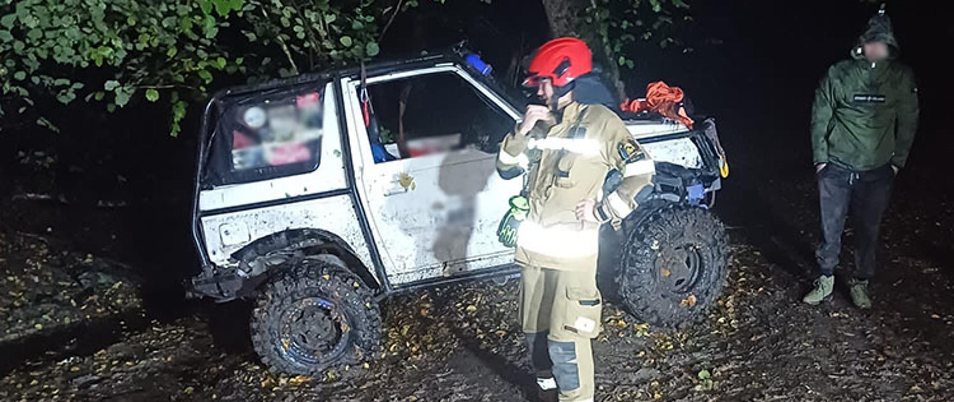 Biały samochód terenowy na tle drzew. Przed pojazdem stoi strażak i rozmawia przez radiotelefon.