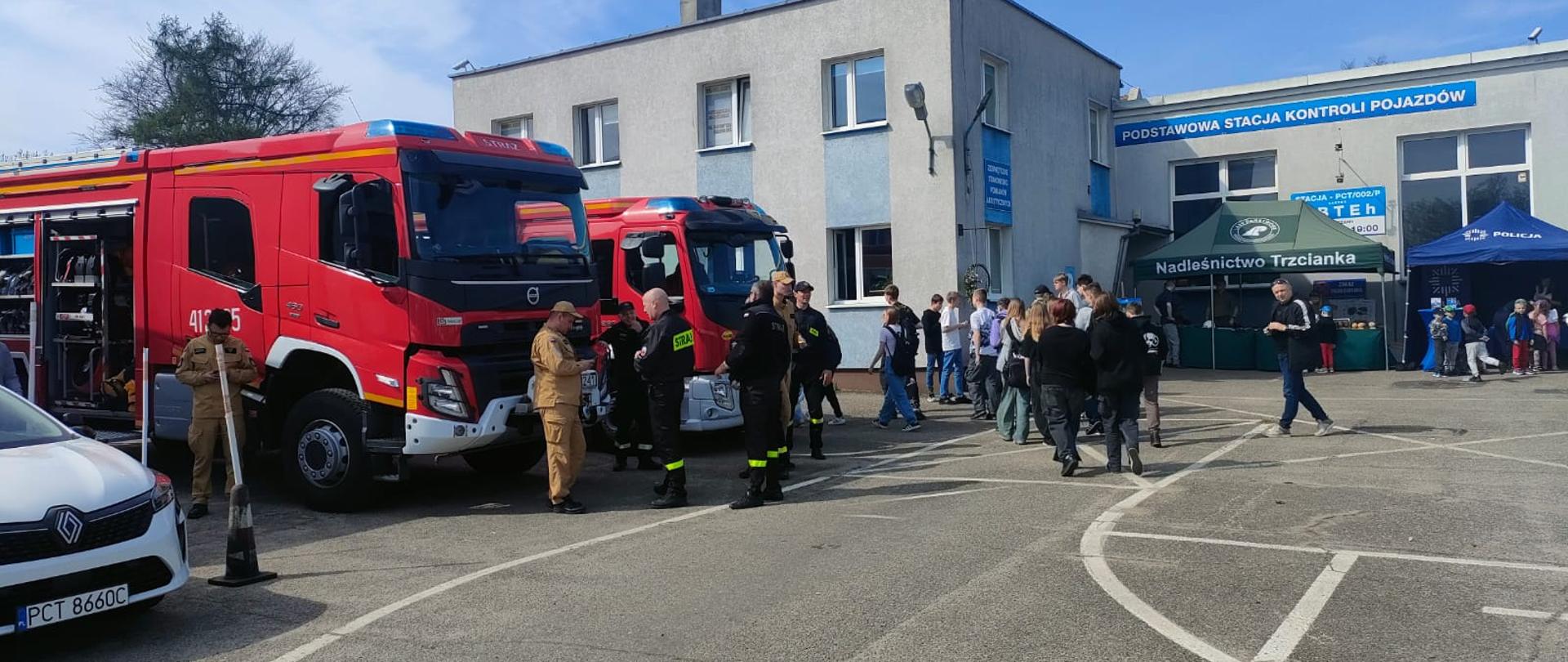Na pierwszym planie widoczne są nowoczesne wozy strażackie z otwartymi przedziałami sprzętowymi. Wokół nich zgromadzeni są funkcjonariusze Państwowej Straży Pożarnej w różnych typach umundurowania oraz odwiedzający – w tym dzieci i młodzież. Po prawej stronie widać namioty promocyjne, m.in. Nadleśnictwa Trzcianka i Policji, przy których odbywa się prezentacja i edukacja.
