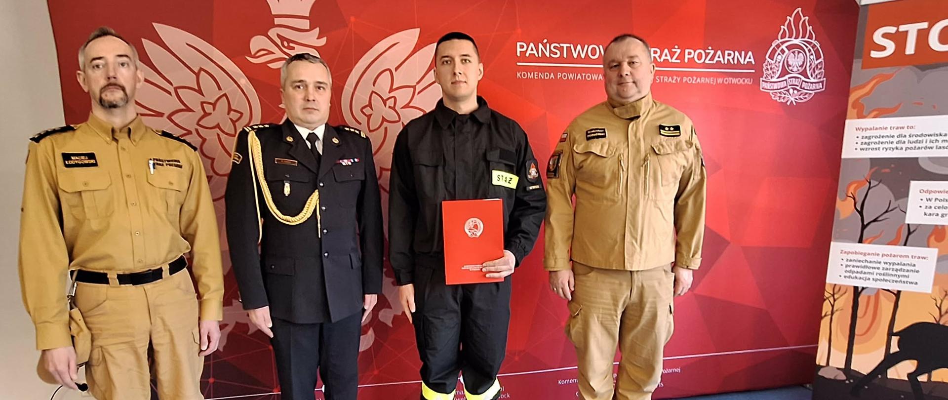 Na zdjęciu widać czterech mężczyzn stojących w pomieszczeniu podczas oficjalnej uroczystości Państwowej Straży Pożarnej. Trzech z nich ma na sobie różne typy umundurowania strażackiego, a jeden — drugi od lewej — występuje w galowym, czarnym mundurze z złotymi akcentami.
Drugi od prawej trzyma w rękach czerwony folder z białym emblematem PSP, co upamietnia wręczenie aktu ślubowania . W tle znajduje się czerwone tło z orłem oraz napisem „Państwowa Straż Pożarna” i informacją o Komendzie Powiatowej PSP w Otwocku. Po prawej stronie stoi roll up z materiałami edukacyjnymi dotyczącymi bezpieczeństwa pożarowego, na którym widnieje m.in. . duży napis „STOP”.
