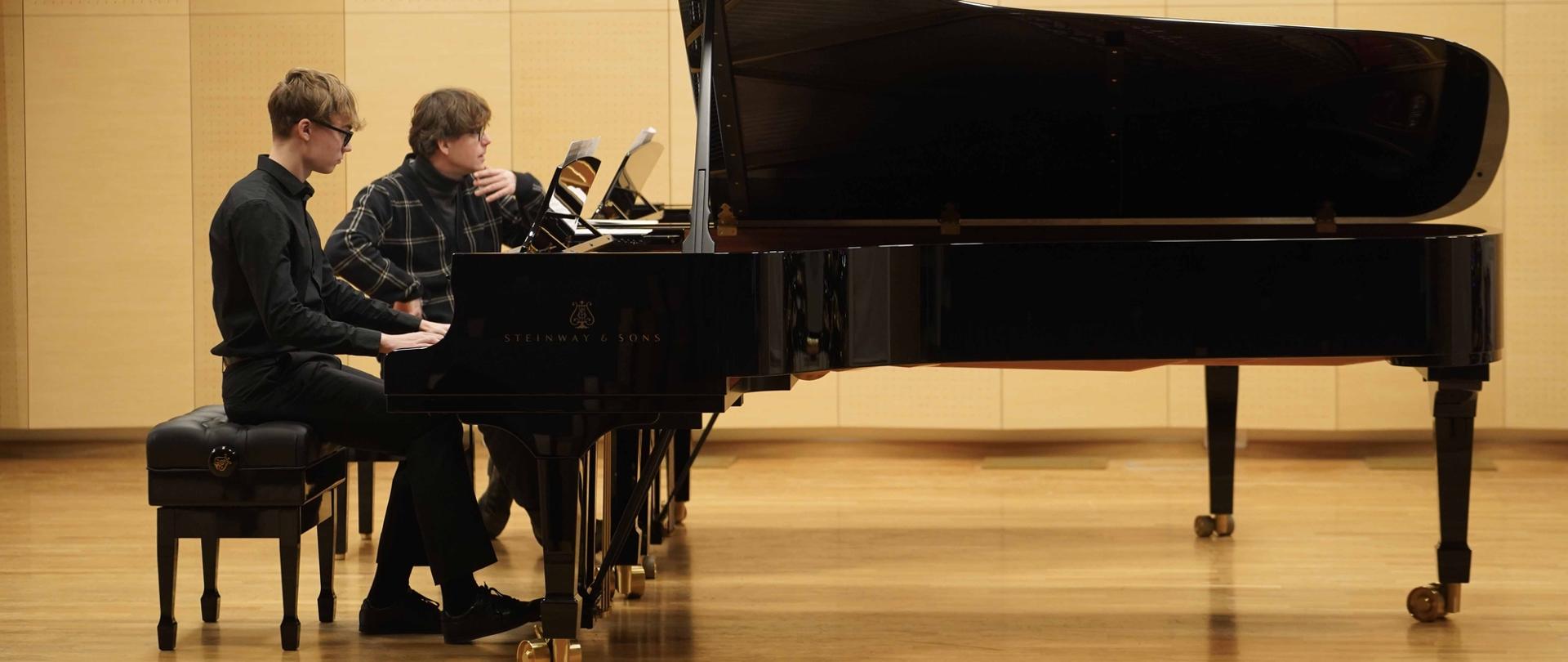 Zdjęcie przedstawia innego ucznia grającego na jednym z fortepianów na scenie sali koncertowej, przy drugim fortepianie prowadzący przysłuchuje się wykonaniu