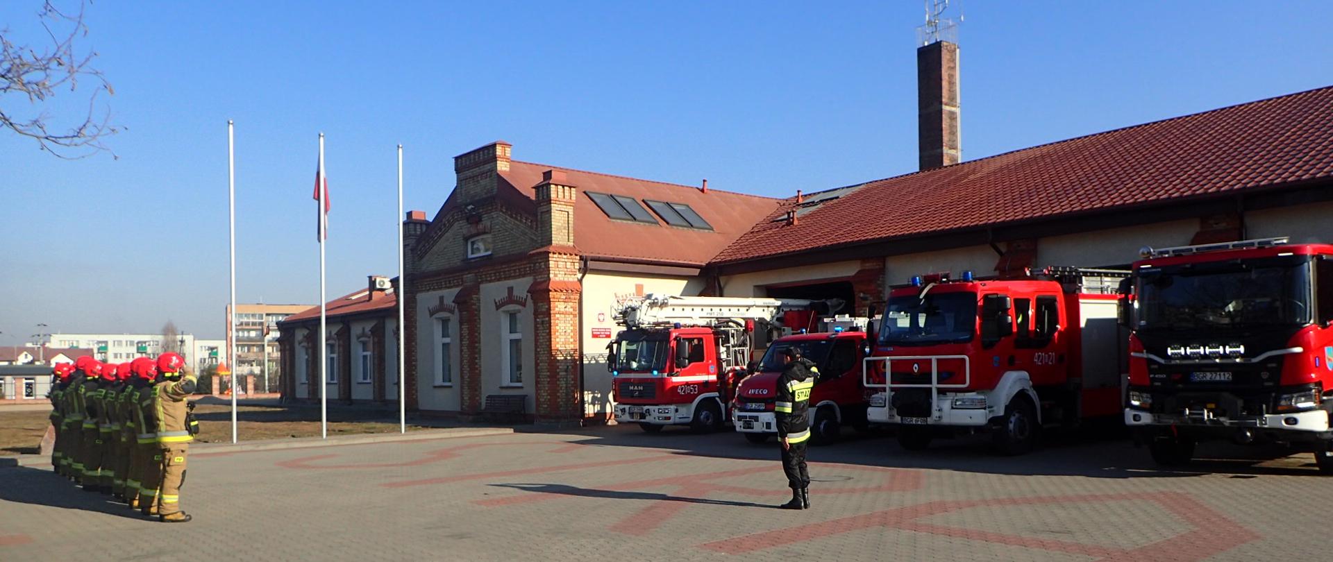Strażacy PSP stoją w rzędzie na placu. Po prawej widoczne pojazdy strażackie stojące przed garażami. Na środku w tle trzy maszty, na jednym powiewa Polska flaga.