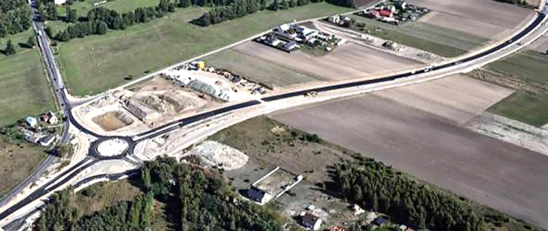Rondo z kilkoma drogami w otoczeniu pól, lasów i budynków, widziane z góry.