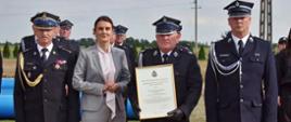 Przedstawicielka władz w szarym garniturze oraz trzej strażacy w galowych mundurach stoją w szeregu i pozują do zdjęcia na świeżym powietrzu. Strażak w środku trzyma w rękach oprawiony w ramkę oficjalny dokument. W tle widać pozostałych uczestników uroczystości.