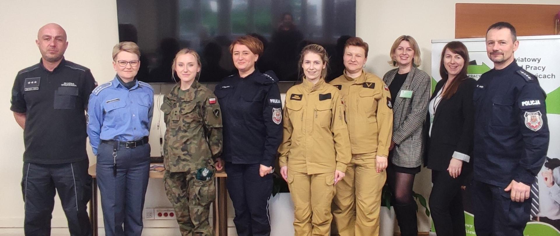 Widok z przodu, przedstawiciele służb mundurowych ustawieni do pamiątkowego zdjęcia.
Od lewej funkcjonariusze służby więziennej, żołnierz Wojska Polskiego, funkcjonariuszka Policji, funkcjonariuszki Państwowej Straży Pożarnej, pracownice Powiatowego Urzędu Pracy oraz funkcjonariusz Policji. 