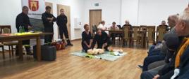 Kobieta ćwiczy zabiegi resuscytacyjne na fantomie. Uciska klatkę piersiową. Strażacy prowadzą instruktarz.