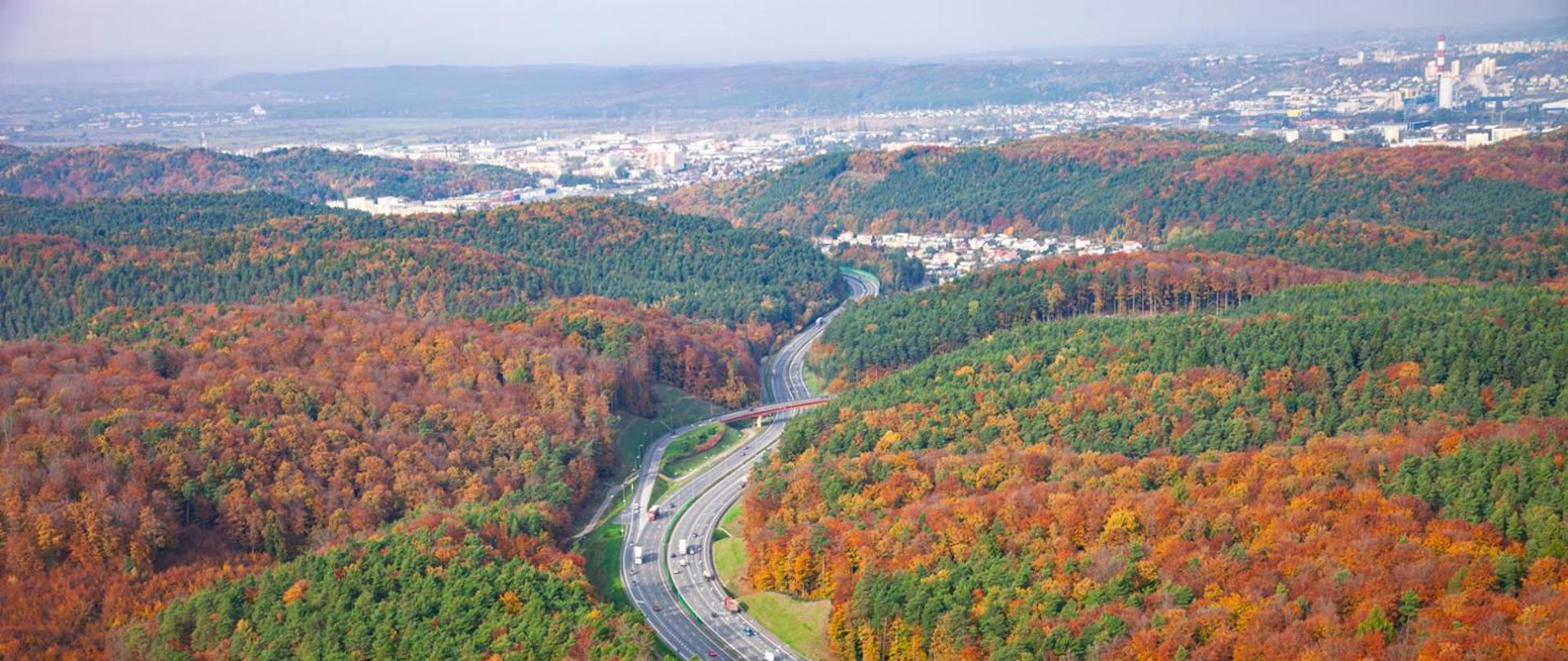 Panorama jesiennego krajobrazu z drogą szybkiego ruchu biegnącą przez las. Wokół widoczne są wzgórza pokryte drzewami w odcieniach zieleni, pomarańczu i czerwieni. W tle rozciąga się miasto z licznymi budynkami i lekką mgłą nad horyzontem.