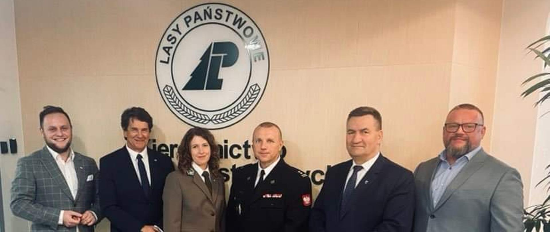 Na zdjęciu sześć osób, przedstawiciele lasów państwowych, samorządu powiatu braniewskiego i komendant powiatowy. Wszyscy stoją na na tle ścianki z logiem lasów państwowych.