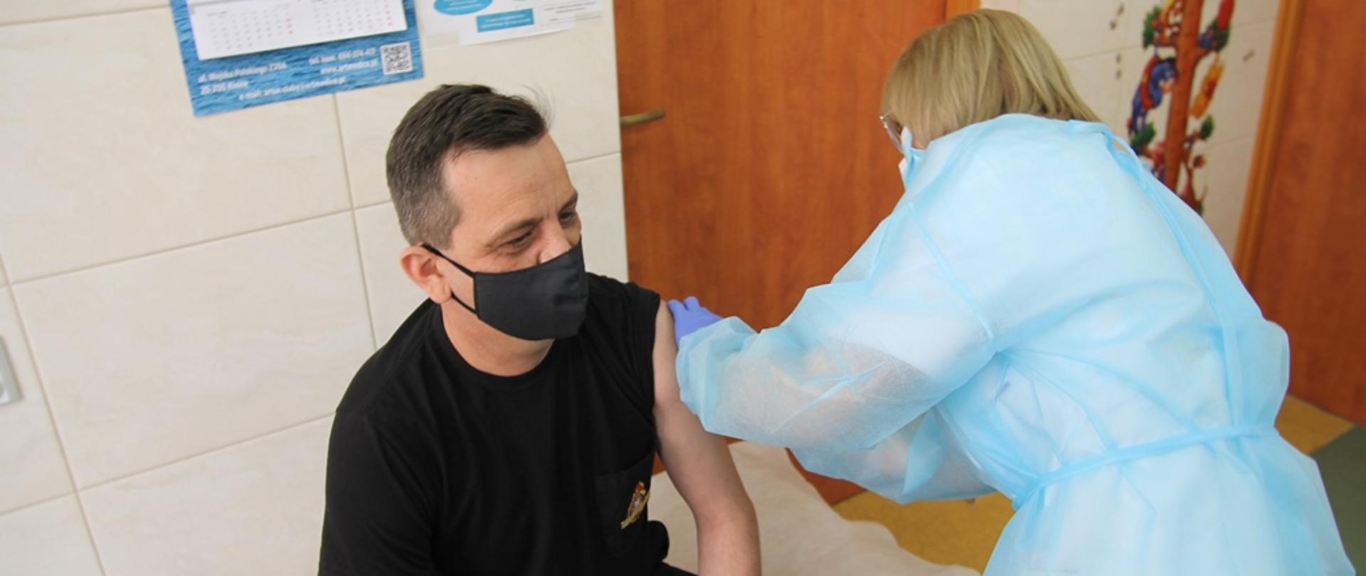 Komendant Powiatowy PSP we Włoszczowie st. kpt. Andrzej Pacanowski podczas szczepienia przez pielęgniarkę w gabinecie zabiegowym. 