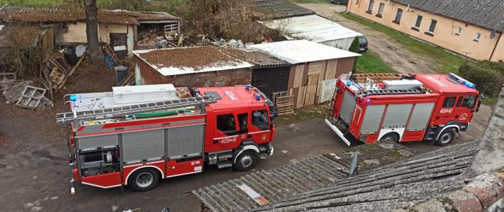 Widok na dwa auta gaśnicze. Zdjęcie wykonane z kosza podnośnika. Auta czerwone, stoją przed budynkiem, w którym prowadzone są działania.