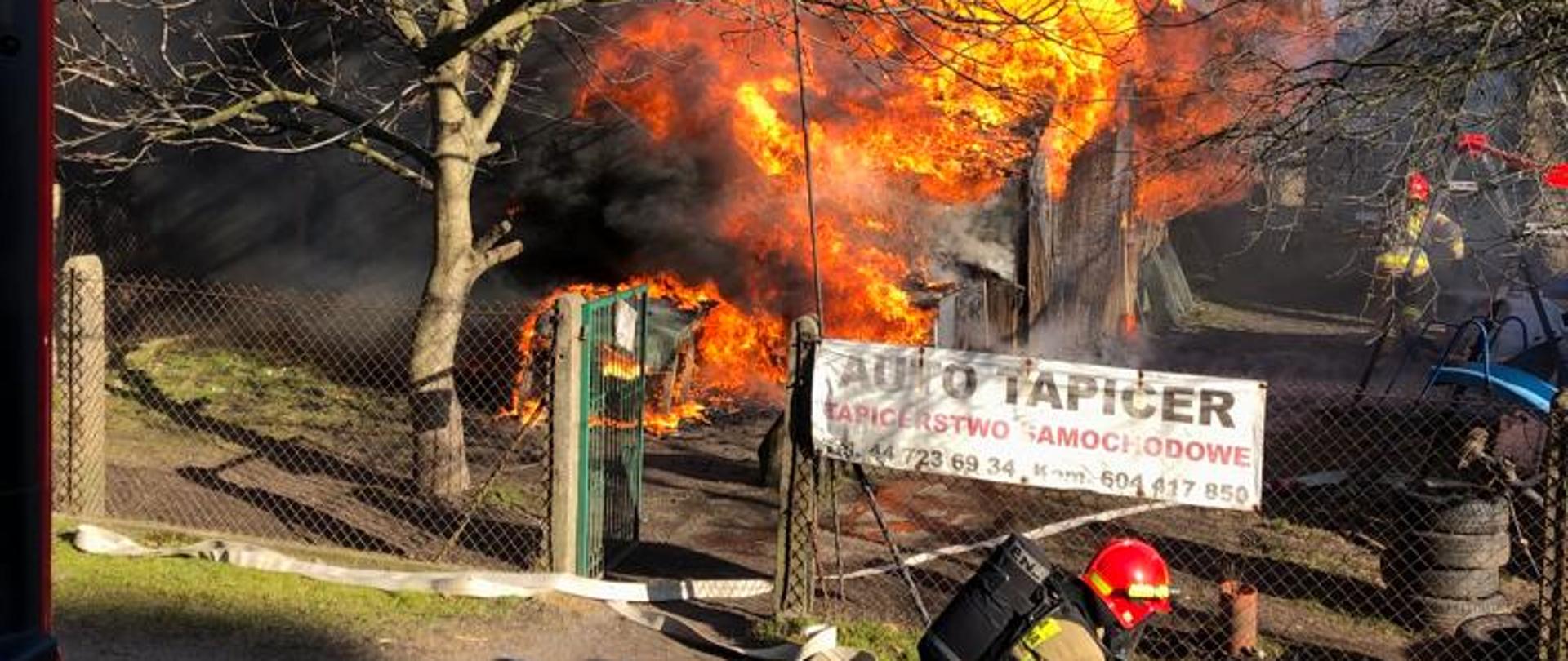 Na pierwszym planie strażak klęczy przy rozdzielaczu. Na drugim planie widać ogień i kłęby czarnego dymu wybijające się w niebo. 