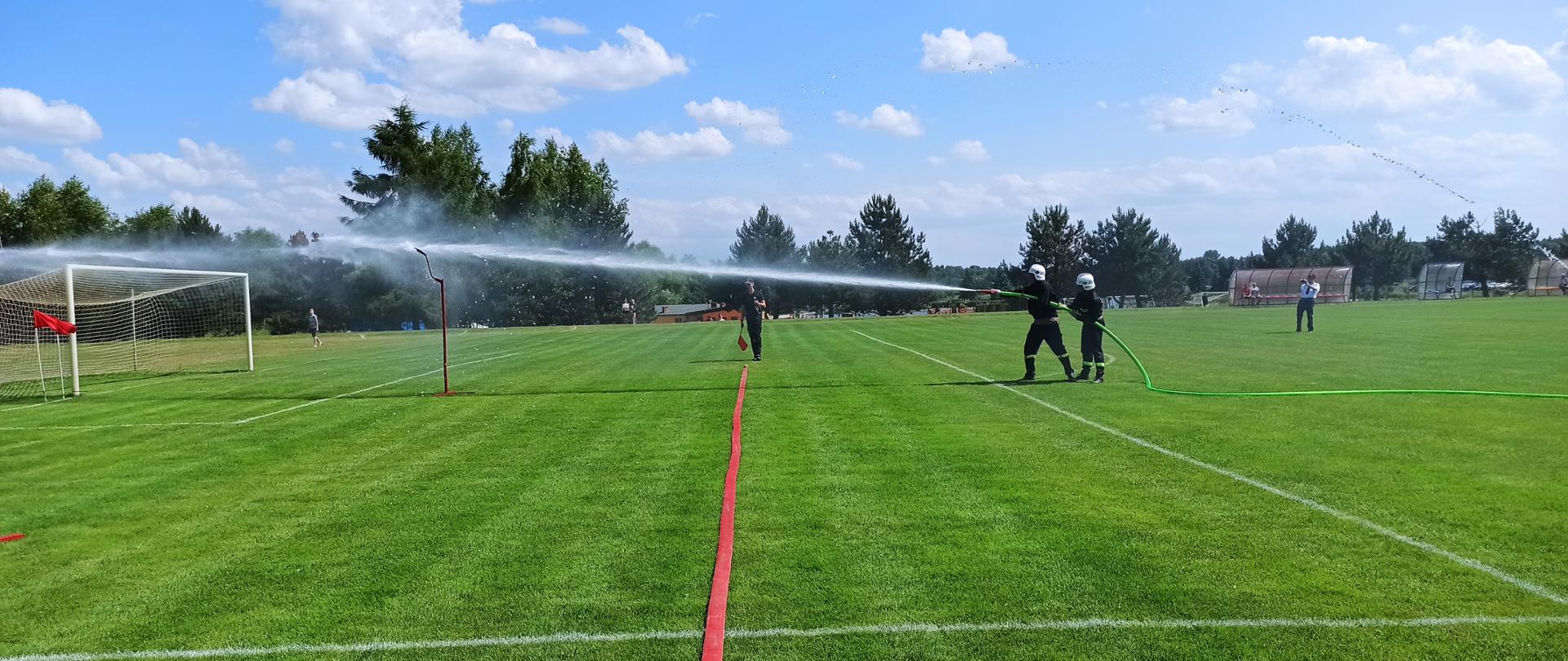 Zdjęcie przedstawia dwóch strażaków stojących jeden za drugim trzymających odcinek wężowy z wodą, celujących w tabliczkę.