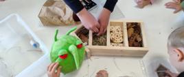 Zdjęcie przedstawia uczestników spotkania w trakcie budowy domków dla zapylaczy w towarzystwie maskotki PESTY
