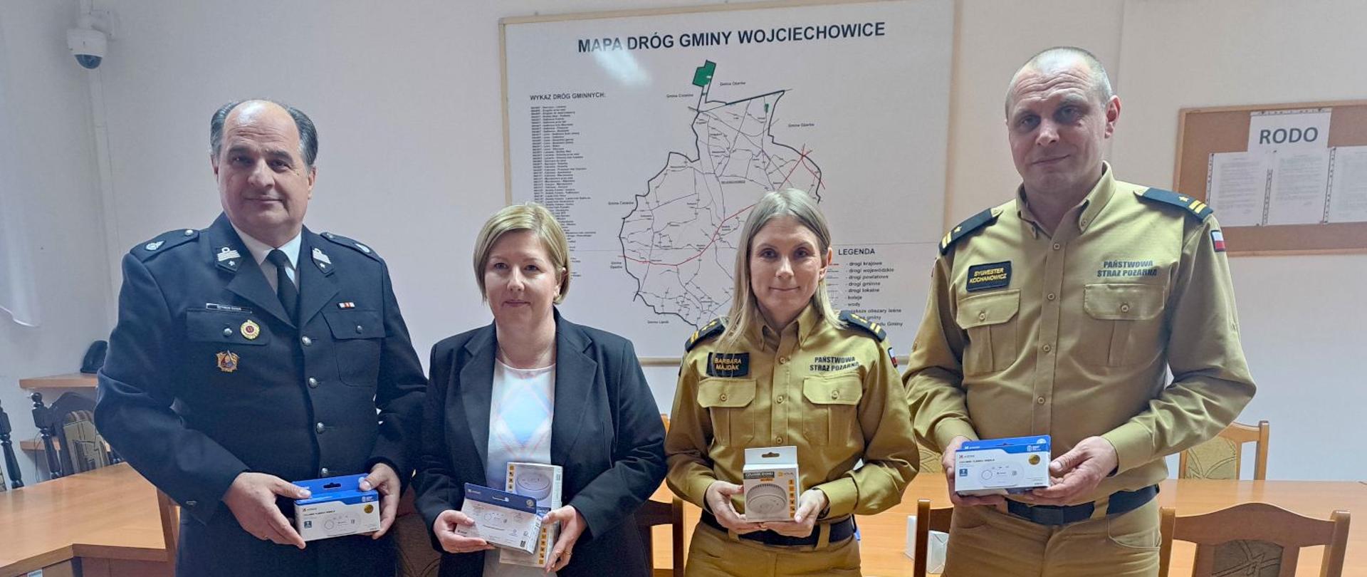 Cztery osoby – dwóch strażaków, wójt gminy i kobieta – stoją w sali urzędowej i prezentują przekazane urządzenia. W tle mapa gminy Wojciechowice