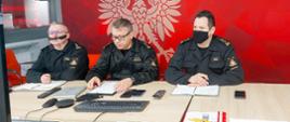 wideokonferencji z kadra kierowniczą PSP woj. mazowieckiego. Strażacy nosza maseczki ochronne. W tle, za plecami strażaków stoi banner z dużym godłem Rzeczypospolitej Polskiej.