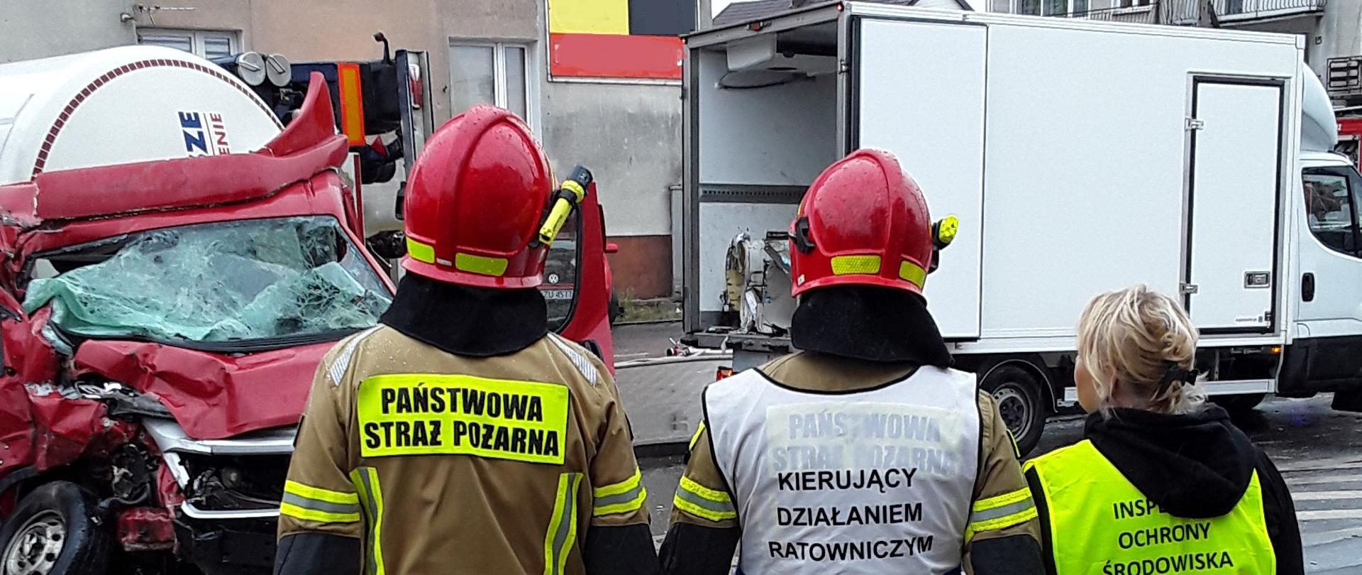 Inspektor Wojewódzkiego Inspektoratu Ochrony Środowiska w Warszawie i dwóch funkcjonariuszy Straży Pożarnej obserwują miejsce kolizji cysterny i auta dostawczego.