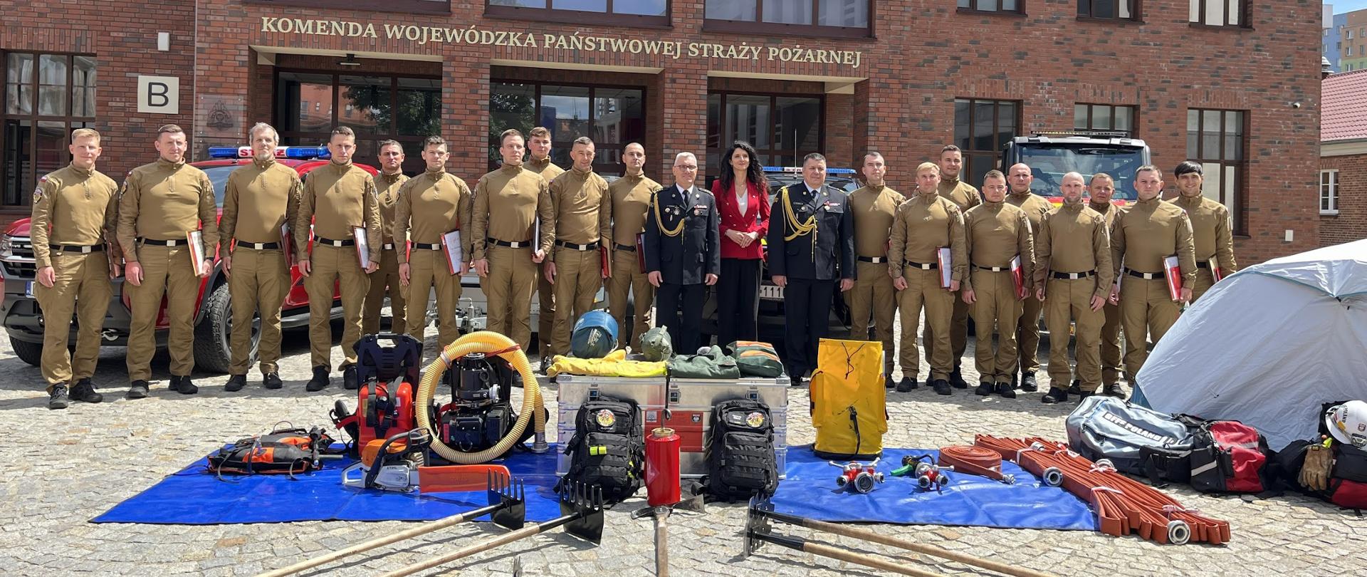 Na zdjęciu znajduje się grupa kilkunastu strażaków w piaskowo-brązowych mundurach polowych oraz trzech oficjeli w ciemnych mundurach galowych i kobieta w czerwonym żakiecie. Wszyscy stoją w rzędzie przed ceglanym budynkiem z dużym, jasnym napisem „KOMENDA WOJEWÓDZKA PAŃSTWOWEJ STRAŻY POŻARNEJ”. Ponad wejściem do obiektu widoczna jest litera „B”.
W tle, za zgromadzonymi osobami, zaparkowane są czerwone samochody strażackie. Na pierwszym planie, na brukowanym placu, rozłożono dwie niebieskie plandeki, na których wyeksponowano specjalistyczny sprzęt ratowniczo-gaśniczy. Wśród wyposażenia widoczne są m.in. motopompy, węże strażackie, torby medyczne, plecaki, hełmy oraz metalowe skrzynie transportowe. Po prawej stronie kadru stoi rozstawiony, jasny namiot bazowy. Bezpośrednio przed plandekami ułożono w wachlarz narzędzia ręczne na długich trzonkach (tłumice). Zdjęcie zostało wykonane w słoneczny dzień.