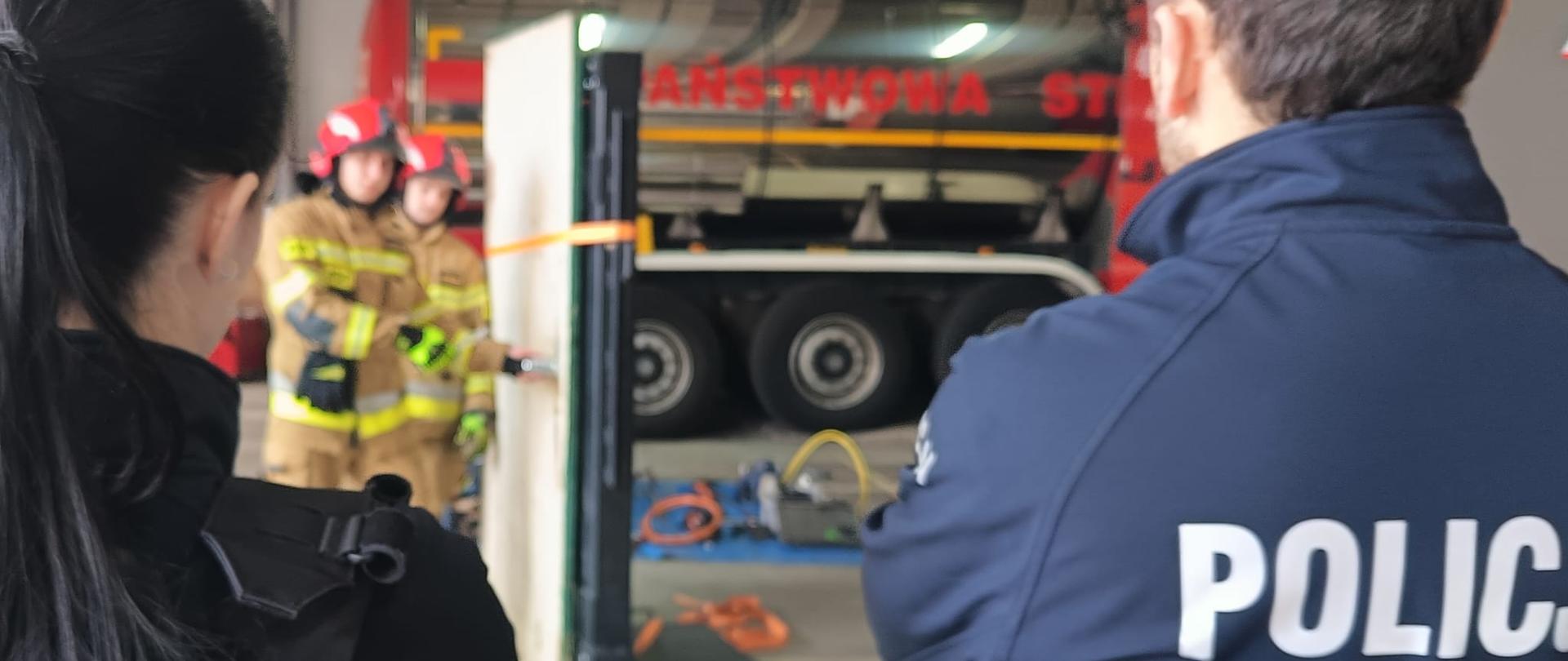 Widok z perspektywy dwóch policjantów stojących tyłem do obiektywu, obserwujących dwóch strażaków przygotowujących się do wyważenia drzwi w remizie strażackiej. Na plecach jednego z policjantów widoczny jest napis "POLICJA". W tle stoi wóz strażacki.