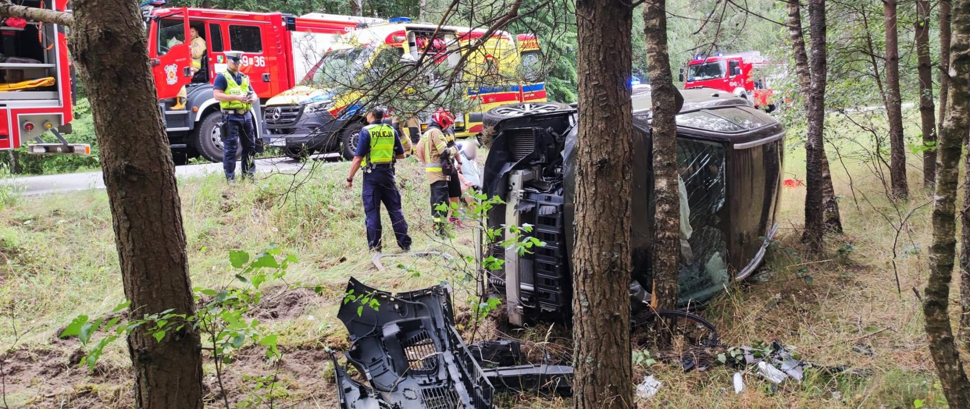 Leżący w rowie na lewym boku samochód osobowy z rozbitym przodem, na drugim planie widać strażaka w czerwonym hełmie oraz ubraniu specjalnym oraz policjantów zwróconych twarzami do siebie, jeden stoi w rowie a drugi na krawędzi jezdni. Za nimi żółta karetka pogotowia oraz trzy czerwone samochody strażackie z włączoną niebieską sygnalizacją świetlną. W samochodzie strażackim z końcówką 36 na miejscu kierowcy przy otwartych drzwiach siedzi strażak w ubraniu specjalnym. 