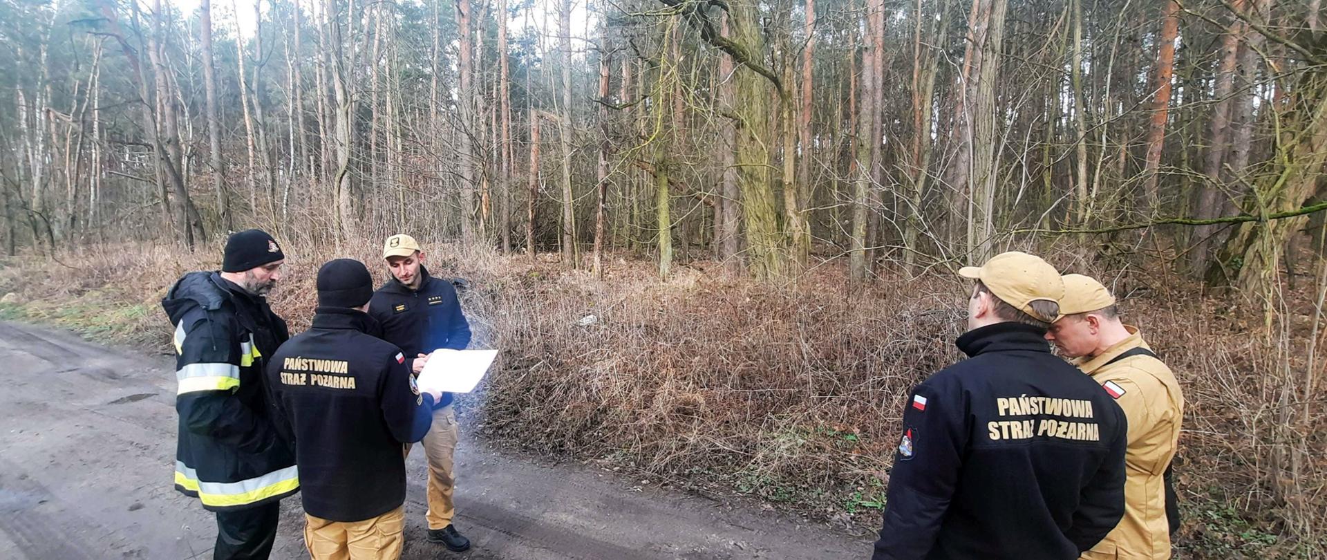 Strażacy stoją na drodze leśnej i omawiają wydrukowany plan.