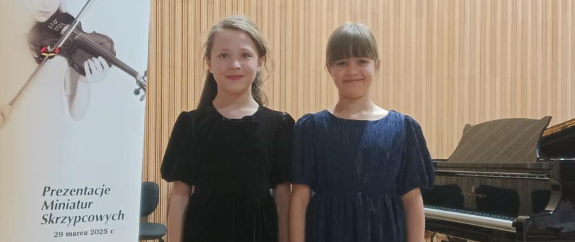 Dwie uczennice klasy skrzypec na scenie koncertowej, w tle fortepian.