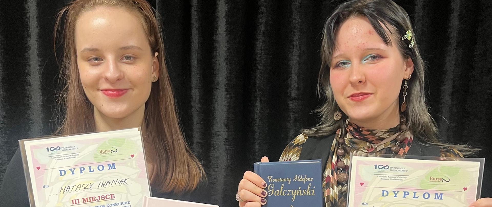 Na tle czarne kurtyny stoją dwie dziewczyny i uśmiechnięte pozują do zdjęcia. Obydwie trzymają w jednej dłoni kolorowy dyplom, a w drugiej książkę.