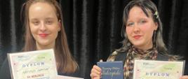 Na tle czarne kurtyny stoją dwie dziewczyny i uśmiechnięte pozują do zdjęcia. Obydwie trzymają w jednej dłoni kolorowy dyplom, a w drugiej książkę.