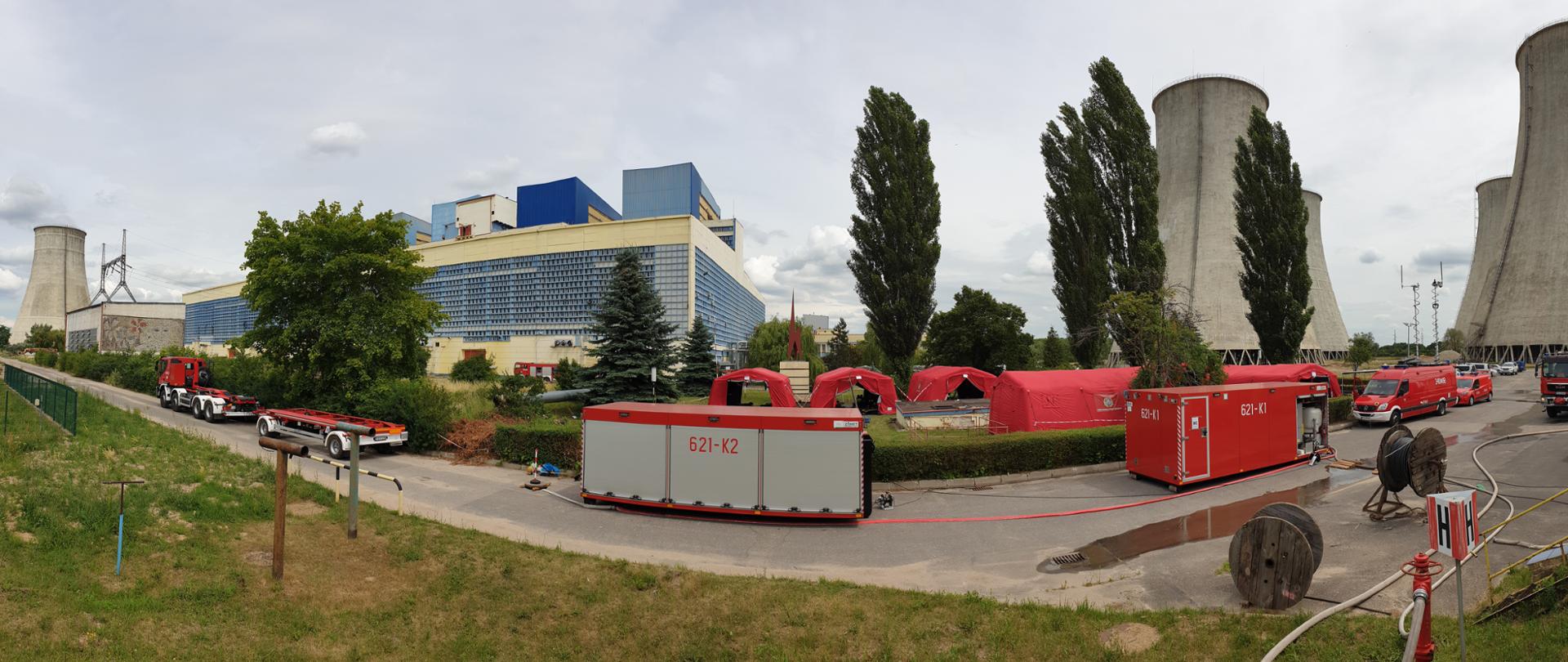 Zdjęcie panorama. Widać zabudowania Elektrowni Adamów w tym chłodnie. Budyenk główny jest w kolorze żółtym, na frontowej ścianie okna z niebieskimi ramami. Na pierwszym planie kontener logistyczny. Po lewej stronie nośnik kontenerów, po prawej samochód dowodzenia i łączności, posiadają kolor czerwony i sygnalizację ostrzegawczą. Za pojazdami namioty sztabu, które są w kolorze czerwonym. Namioty znajdują się na pasie zieleni pomiędzy drogami. W prawym rogu czerwony hydrant i rozwinięte od niego węże strażackie w kolorze szarym. Niebo zachmurzone, w kolorze szarym.
