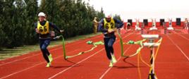 Zbliżenie na dwóch zawodników PSP w trakcie konkurencji 100 m z przeszkodami. W rękach węże pożarnicze, końcówka biegu na torze z czerwonego tartanu.